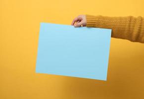 woman in an orange sweater holds a blank sheet of paper on a yellow background. Place for an inscription, advertisement, information photo