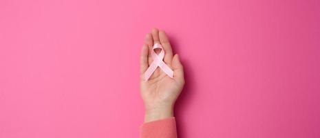 female hand hold a pink silk ribbon in the form of a loop on a pink background. Symbol of the fight against breast cancer