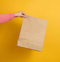 woman holding in hand brown blank craft paper bag for takeaway on yellow background. Packaging template mock up. Delivery service concept photo