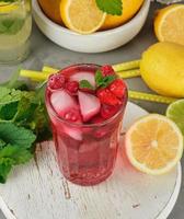 red strawberry lemonade in a glass on a round white wooden board, yellow lemons are behind photo