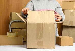 woman in a gray sweater is packing brown cardboard boxes on a white table, behind a stack of boxes photo