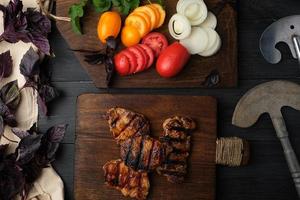 trozos de carne de cerdo a la parrilla, junto a una tabla con verduras frescas picadas foto