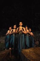 A Bunch of girls in green costumes holding a wooden masks in their hands while standing between the big rocks in front of the forest photo