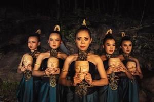 A Bunch of girls in green costumes holding a wooden masks in their hands while standing between the big rocks in front of the forest photo