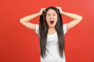 retrato de una joven asiática frustrada y enojada con una camiseta blanca tirando de su cabello aislada de fondo rojo