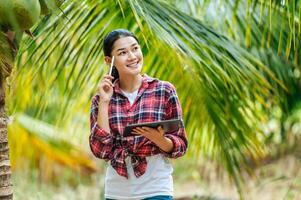 Portrait of Happy Asian young  farmer woman check quality of coconut in farm and thinking while standing and holding tablet. Agricultural and technology concepts. photo