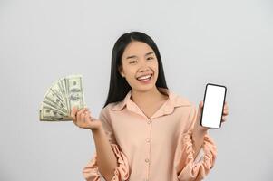 retrato de una joven alegre sosteniendo billetes de dinero y mostrando una maqueta de smartphone aislada sobre fondo blanco foto