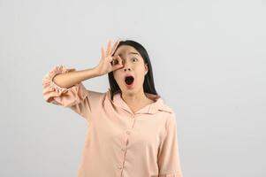 Portrait of Excited young asian woman in pink shirt doing ok hand over face isolated on white background photo