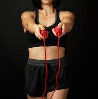 mujer joven con una figura deportiva en uniforme negro sostiene una cuerda roja para saltar foto