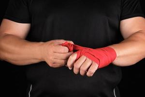 athlete stand in black clothes and wrap his hands in red textile elastic bandage before training photo