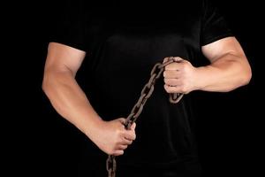 adult man in black clothes stands upright with strained muscles and holds a rusty metal chain photo