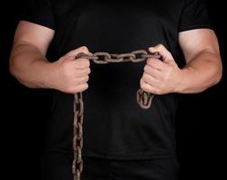 adult man in black clothes stands upright with strained muscles and holds a rusty metal chain photo