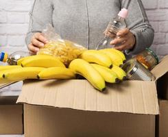 mujer con suéter gris pone en una caja de cartón varios alimentos, frutas, pasta, aceite de girasol en una botella de plástico y conservas foto