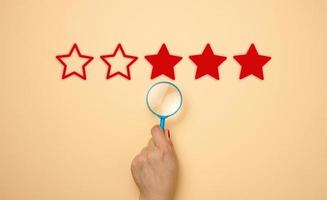 five stars and a hand with a blue plastic magnifier on a beige background. Evaluation of the quality of services and goods, high rating photo