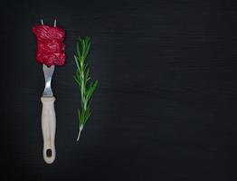 raw pieces of meat on an iron kitchen fork and a green branch of rosemary photo