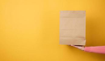 woman holding in hand brown blank craft paper bag for takeaway on yellow background. Packaging template mock up. Delivery service concept photo