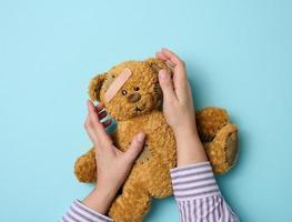 female hand holds a brown teddy bear and glues a medical adhesive plaster on a blue background, tram treatment photo