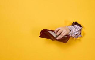 A woman's hand holds an empty leather wallet, part of the body sticks out of a hole with torn edges photo