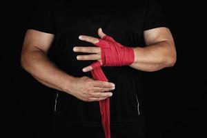 athlete stand in black clothes and wrap his hands in red textile elastic bandage before training photo