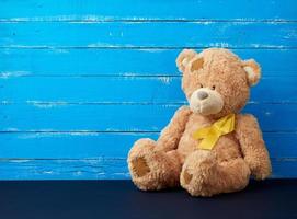 brown teddy bear sits and a yellow silk ribbon on a blue wooden background, concept of the fight against childhood cancer photo