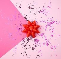 red bow and shiny multi-colored round confetti scattered on a pink background photo