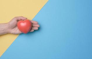 Female hand holds red heart, blue yellow background. Love and donation concept, top view photo