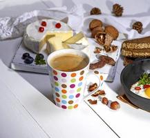 coffee with brown foam in a white ceramic mug, behind a round camembert cheese photo