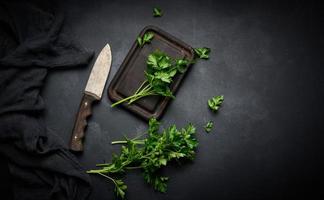 ramitas verdes de perejil en una tabla de cortar de madera y un cuchillo. vista superior de la mesa de la cocina negra foto