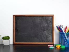 pizarra negra vacía, lápices multicolores en un soporte de metal azul sobre una mesa negra foto