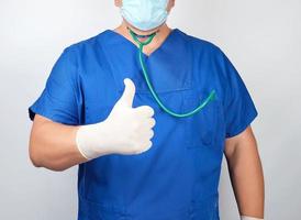 médico masculino con uniforme azul y guantes blancos de látex muestra un gesto con la mano derecha como