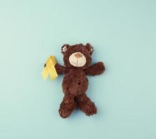 brown teddy bear holds in his paw a yellow ribbon folded in a loop on a blue background. concept of the fight against childhood cancer photo