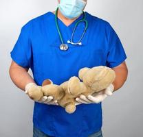 médico con uniforme azul y guantes de látex blancos sosteniendo un oso de peluche marrón foto