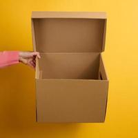 female hand holding an open brown cardboard box on a yellow background, delivery photo