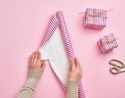 two female hands unfold a roll of wrapping paper, next to two gifts, pink background photo