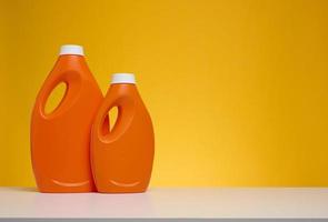 dos botellas de plástico naranja con detergente líquido sobre una mesa blanca, fondo amarillo foto