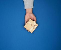 female hand in a gray sweater holds a closed square cardboard gift box with a bow photo