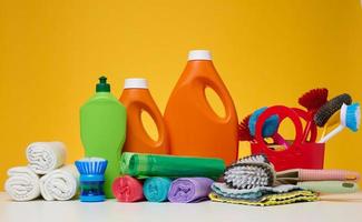 botellas de plástico naranja con productos líquidos y bolsas de basura, cepillos y trapos textiles para lavar suelos sobre una mesa blanca foto