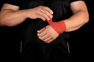 athlete stand in black clothes and wrap his hands in red textile elastic bandage photo