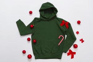 Close up green blank template hoodie copy space. Christmas Holiday concept. Top view mockup hoodie. Red holidays decorations on jumper white background. Happy New Year accessories. Selective focus photo