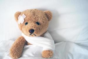 oso de peluche acostado enfermo en la cama con una diadema y un paño cubierto foto