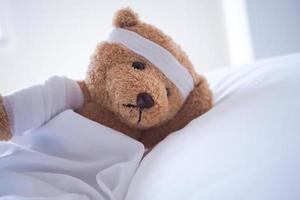 Teddy bear lying sick in bed With a headband and a cloth covered photo