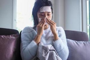 Sick woman sitting on the sofa in the house, having high fever on the forehead with a gel pad to reduce body temperature, sore throat, runny nose and coughing. photo