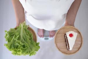 Women weigh themselves on scales and holding vegetables and chocolate cakes. Vegetables are useful to the body and the cake has trans fat. diet concept photo