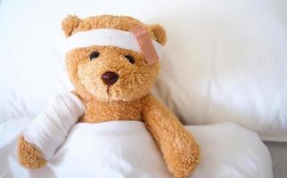 oso de peluche acostado enfermo en la cama con una diadema y un paño cubierto foto