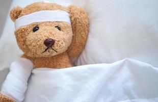 oso de peluche acostado enfermo en la cama con una diadema y un paño cubierto foto