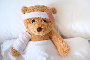 Teddy bear is sick on the bed, his arm is broken and his head is broken from an accident. photo