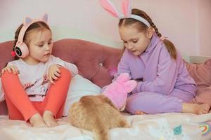 two sister girls play on the bed with a ginger domestic cat in an Easter hat, listen to music on headphones, the older girl has a headband with bunny ears on her head, family preparation for Easter photo