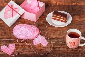 On a wooden background are knitted pink homemade hearts, knitting tools, yarn, a souvenir box and a dessert with tea. Valentine's day, manual labor, hobby photo