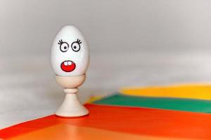 on colored paper, a stand with a white egg, on which there is a sticker with the emotion of surprise, fright, minimalism, copy space. communication on the Internet using emoticons with emotions photo