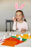 a little girl in bunny ears on her head decorates white eggs with stickers with different emotions, in her hands is a white egg with emotion, the child copies and shows with facial expressions. photo